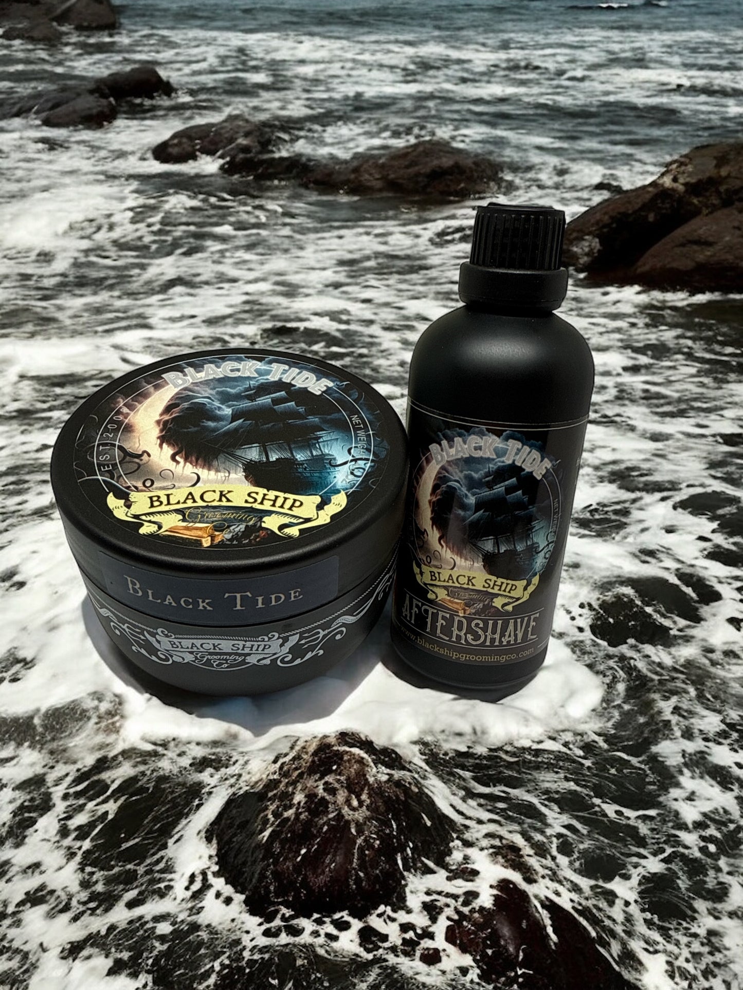 bottle of 'Black Tide' aftershave  and shaving soap by 'Black Ship grooming co.' on a rocky beach with ocean waves.