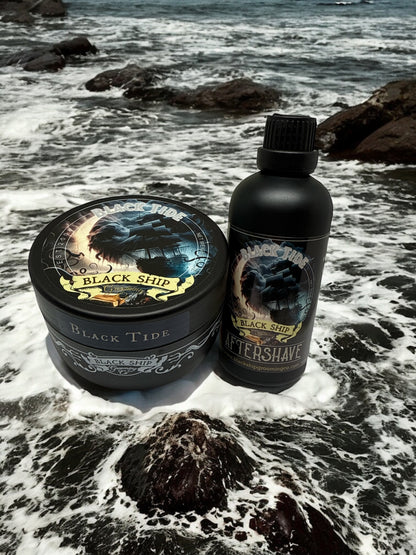 bottle of 'Black Tide' aftershave  and shaving soap by 'Black Ship grooming co.' on a rocky beach with ocean waves.